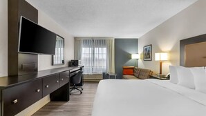 Quarto de hotel com uma cama, escrivaninha, cadeira, televisão, luminária e janela com cortinas.