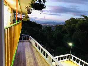 Un balcón con piso de madera y barandal blanco, con vista a una ciudad y montañas.
