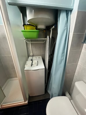 Et lille badeværelse med toilet i hvid farve, en vaskemaskine, et grønt håndvask på en hylde og en bruseforhæng.