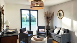 Un salon moderne avec des meubles en osier, une table basse en verre et une vue sur un jardin, accessible par une porte-fenêtre coulissante.