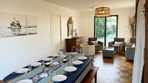 Un coin repas avec une longue table dressée pour six personnes, entourée de chaises en osier, d’un canapé et d’une grande fenêtre donnant sur un paysage verdoyant.