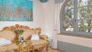 A room with a gold ornate sofa, a painting on the wall, and a large arched window with a view of trees.