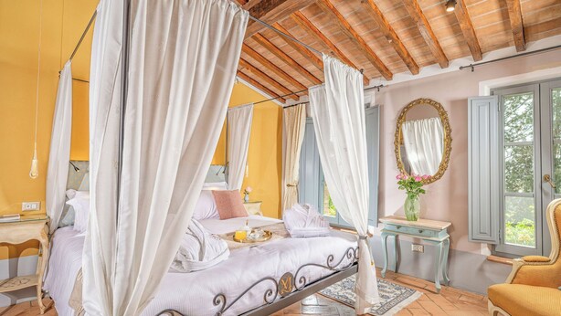A four-poster canopy bed with white linens, a wooden ceiling, a mirror, and a window with shutters.