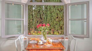 Ein Esstisch mit Tellern, Tassen und einer Blumenvase, vor einem Fenster mit Blick auf einen Hecken- und Baumgarten.