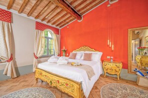 Ein Schlafzimmer mit einem großen Bett, einem hölzernen Kopfteil und einer roten Wand. Es gibt ein Fenster mit Vorhängen, einen Nachttisch mit einer Vase und einen Spiegel an der Wand.