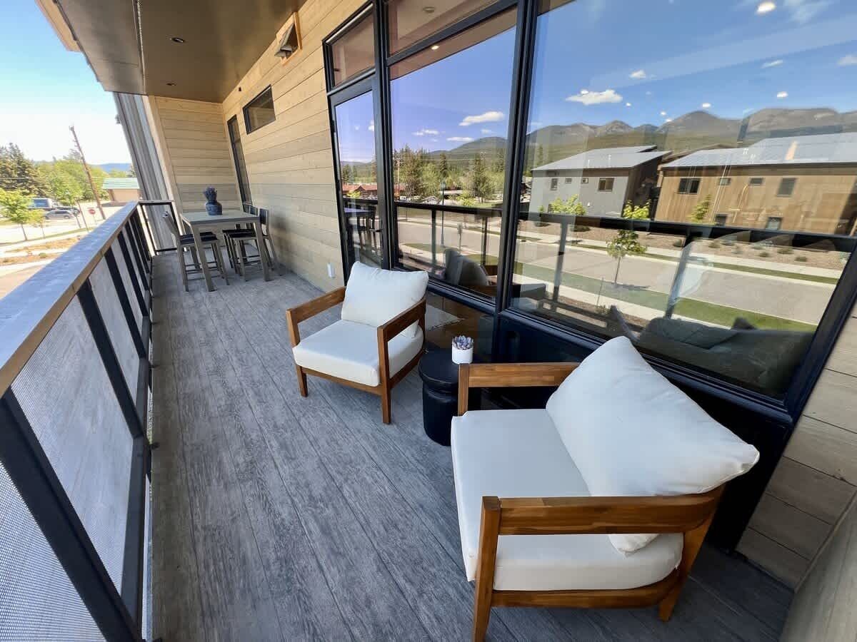 Een balkon met twee houten stoelen en een tafel, grote ramen bieden uitzicht op gebouwen en bergen.