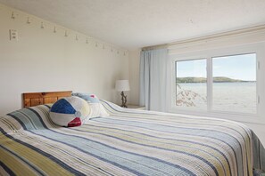 Ein Hotelzimmer mit Bett, Nachttisch, Lampe, Fenster mit Meerblick und Wand mit Steckdosen.