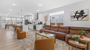 Uma sala de estar moderna com um sofá de couro marrom, uma mesa de centro de vidro e uma poltrona marrom. Dá para ver uma cozinha ao fundo, com armários brancos e uma área de jantar com um banco.