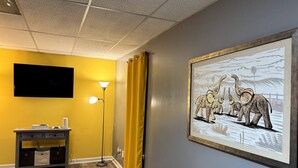 A room with a flat-screen TV, a console table with a speaker, a floor lamp, and a framed picture of elephants. - Quiet, charming 4-bedroom house; 20mins from Atlanta & Airport. (Lithia Springs)