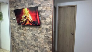 A room with a stone wall, a flat-screen TV displaying a fireplace, a wooden door, and a tiled floor. - BAM - Lodging (Santa Marta)