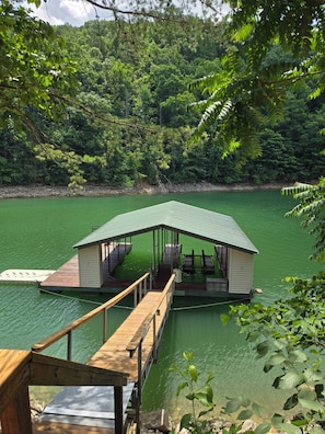 A wooden dock leading to a small cabin with a green roof, surrounded by dense greenery and a calm lake. - Lakefront - Norris Lake - Hot Tub, 2 Slip Covered Dock, Kayaks, ATV trails (Jacksboro)