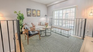 A cozy corner with a leather sofa, a wooden chair, a round table, and a bench. There are three framed pictures on the wall, a large window with a view, and a potted plant. - Perfect Traveling Nurse Housing - Theatre District (Boston)