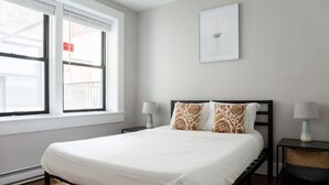 Une chambre à coucher avec un lit, deux tables de chevet avec des lampes, une fenêtre avec des stores et un tableau encadré au mur.