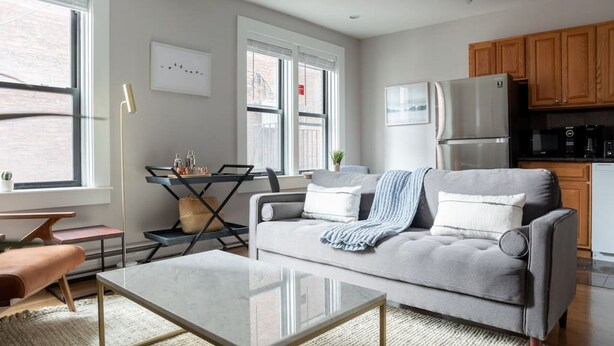 Un salon moderne avec un canapé gris, une table basse en verre et un coin cuisine doté de meubles en bois et d’un réfrigérateur en acier inoxydable.