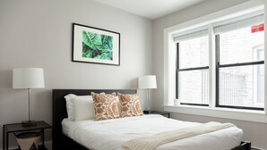 A modern bedroom with a large bed, two bedside tables with lamps, a framed picture on the wall, and a window with blinds.