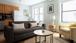 A modern living room with a dark grey sofa, a round coffee table, and a beige armchair. There is a kitchen area with wooden cabinets and a refrigerator.