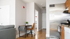A modern kitchen with stainless steel appliances, a dining area with a grey sofa and a small table, and a hallway leading to a door. - Airy 1BR Walkable to Everywhere in Boston | W+D (Boston)