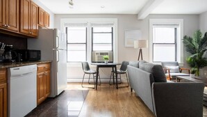 A modern kitchen with wooden cabinets, a dishwasher, and a dining area with a gray sofa and chairs. - Airy 1BR Walkable to Everywhere in Boston | W+D (Boston)