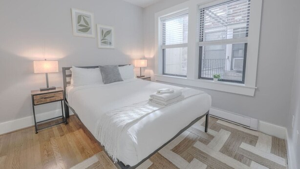 A bedroom with a bed, two bedside tables with lamps, a window with blinds, and a small plant on the windowsill.