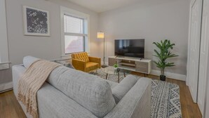 A living room with a grey sofa, a yellow armchair, a flat-screen TV, a small table, and a potted plant.