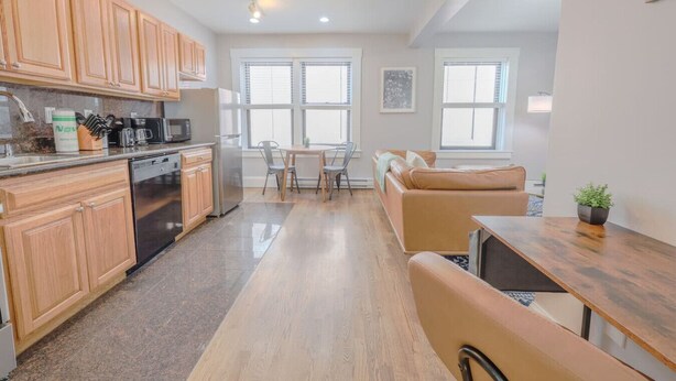 A modern kitchen with wooden cabinets, a dining area with a table and chairs, and a sofa in the living area. - Bay Village Charming 1 Bedroom Apartment (Boston)
