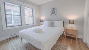 Ein Schlafzimmer mit einem großen Bett, zwei Nachttischlampen, einem gerahmten Bild an der Wand und einem Fenster mit Blick auf ein Backsteingebäude.
