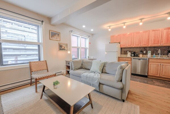A modern living room with a sofa, a coffee table, and a dining table. There is a kitchen area with wooden cabinets and a dishwasher. - Chic 1 Bed Apartment! | Steps from Boston Common! (Boston)