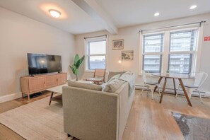 Una sala de estar moderna con un televisor de pantalla plana, un sofá, una mesa de comedor con sillas y ventanas amplias con persianas.