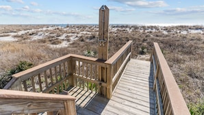 Una terraza de madera con barandilla da a una playa con vegetación escasa y un cielo despejado.