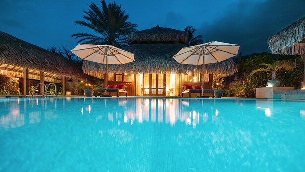 Ein Pool mit Liegestühlen und Sonnenschirmen, umgeben von strohgedeckten Gebäuden und Palmen.