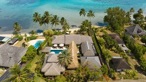 Ein Luftbild eines tropischen Resorts mit strohgedeckten Gebäuden, einem Swimmingpool und einem Strand mit klarem, türkisblauem Wasser.