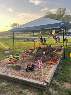 A sandy play area with a tent overhead, surrounded by grass and trees. - Cool Custom Cabin Right Next to Pool! (Claremore)