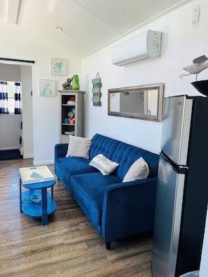 Una sala moderna con un sofá azul, una pequeña mesita auxiliar, un refrigerador y un aire acondicionado instalado en la pared.