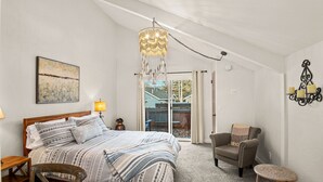 Quarto com cama, uma cadeira, uma mesinha, um candelabro e uma porta de vidro corrediça que dá para uma área externa.
