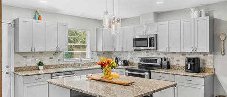 A modern kitchen with white cabinets, granite countertops, a central island with stools, a microwave, stove, and a hanging light fixture.