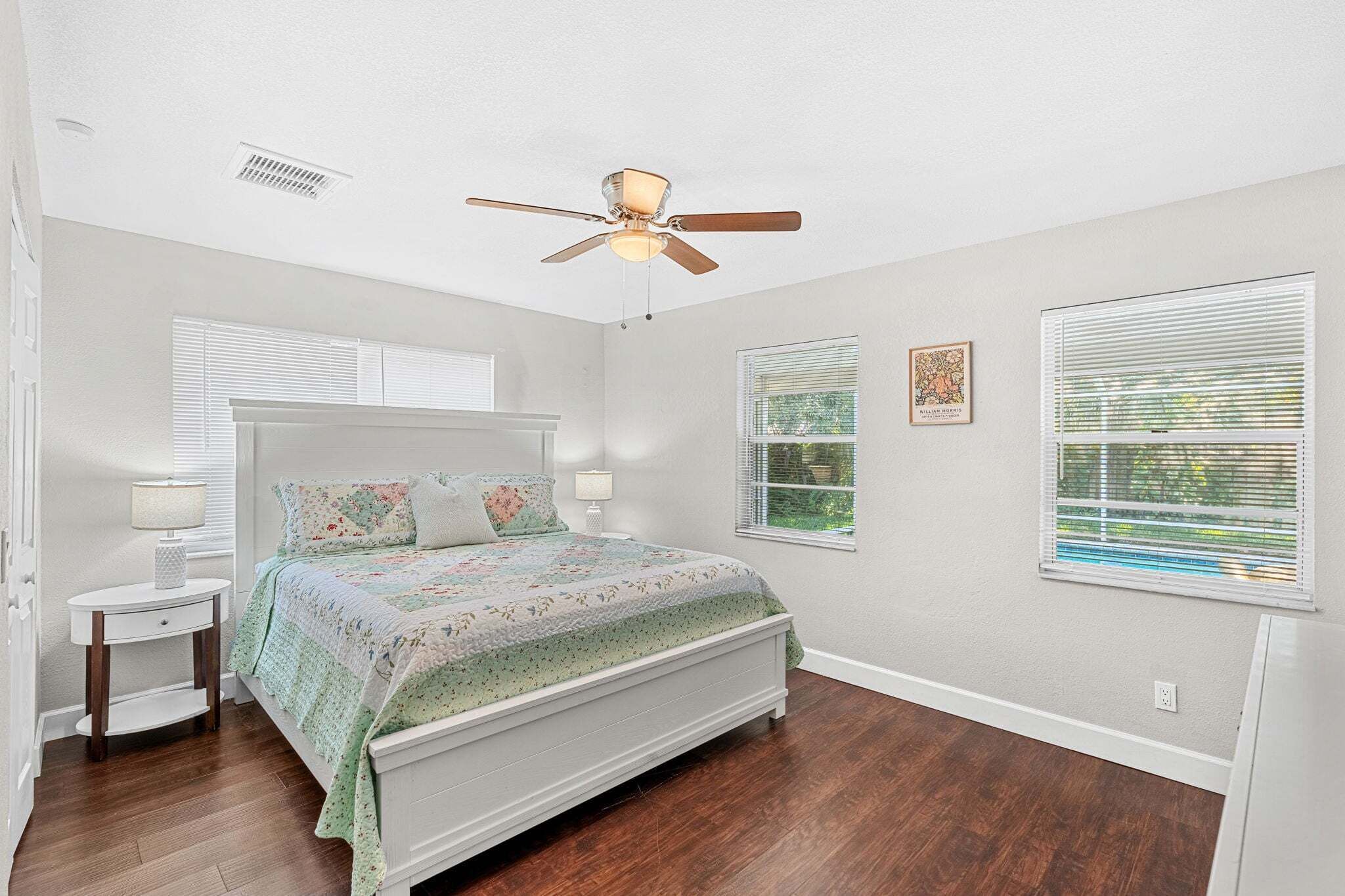 A bedroom with a bed, a ceiling fan, two windows with blinds, a nightstand, and a wall-mounted light.