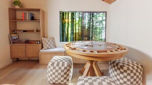 Ein gemütliches Zimmer mit einem runden Holztisch, einem Bücherregal, einer gepolsterten Bank und einem großen Fenster mit Blick auf einen Wald.