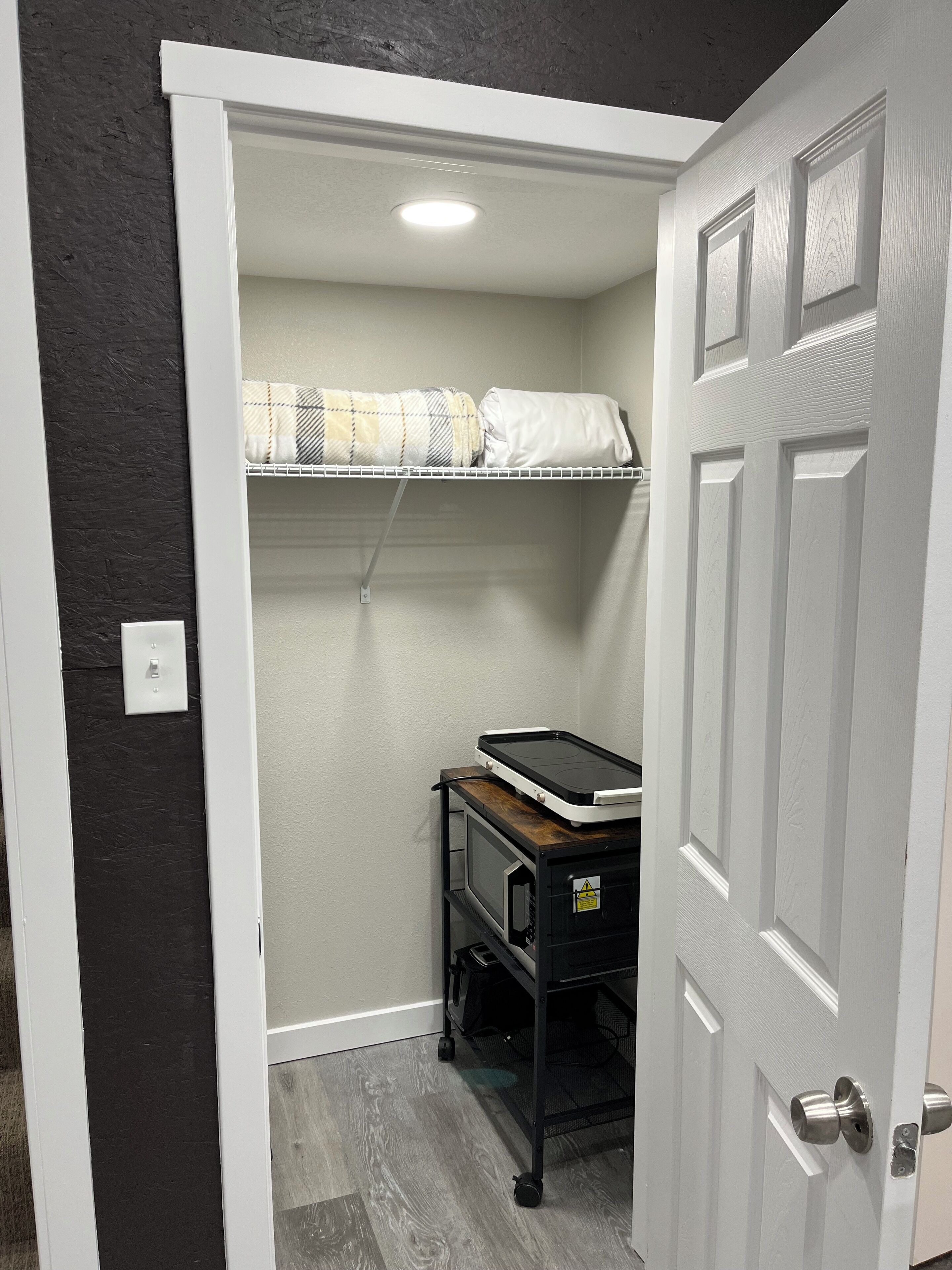 A compact room with a white door, a small shelf with a microwave, and a bed with a checkered blanket.