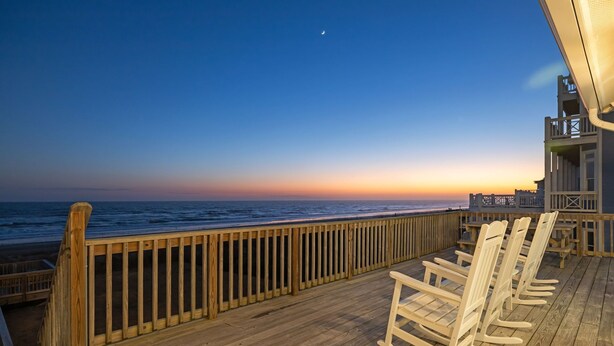 Une terrasse en bois avec des fauteuils à bascule donnant sur l’océan au coucher du soleil.