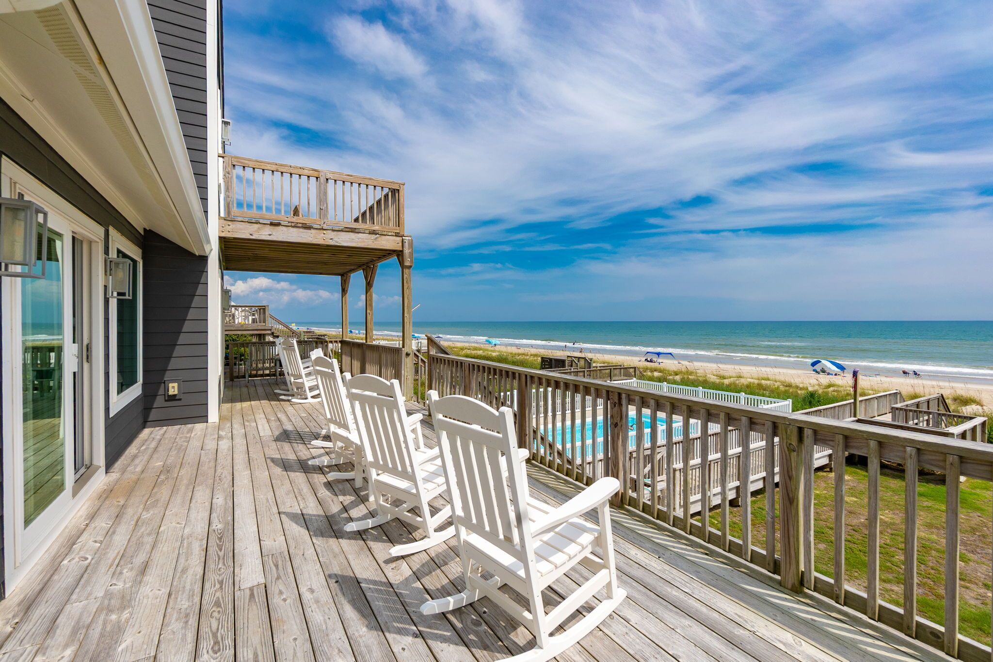 A wooden deck with white rocking chairs overlooking a beach and a swimming pool.