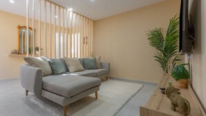 Ein Wohnzimmer mit Ecksofa, einem weißen Teppich, einem Holztisch, einem an der Wand montierten Fernseher und einer Topfpflanze.