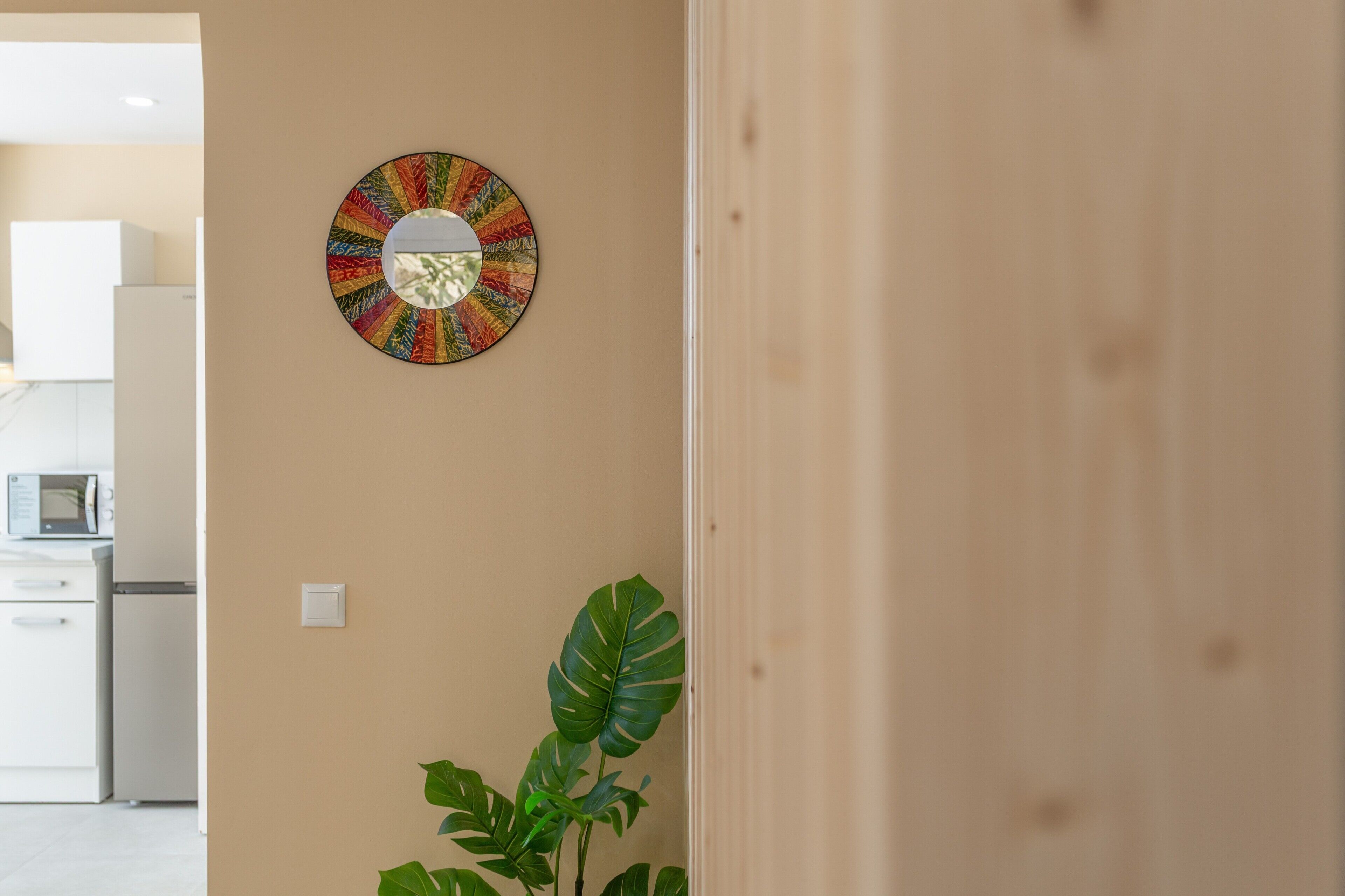 Una habitación con un espejo en la pared, una planta en maceta y una cocina visible a través de una puerta abierta.