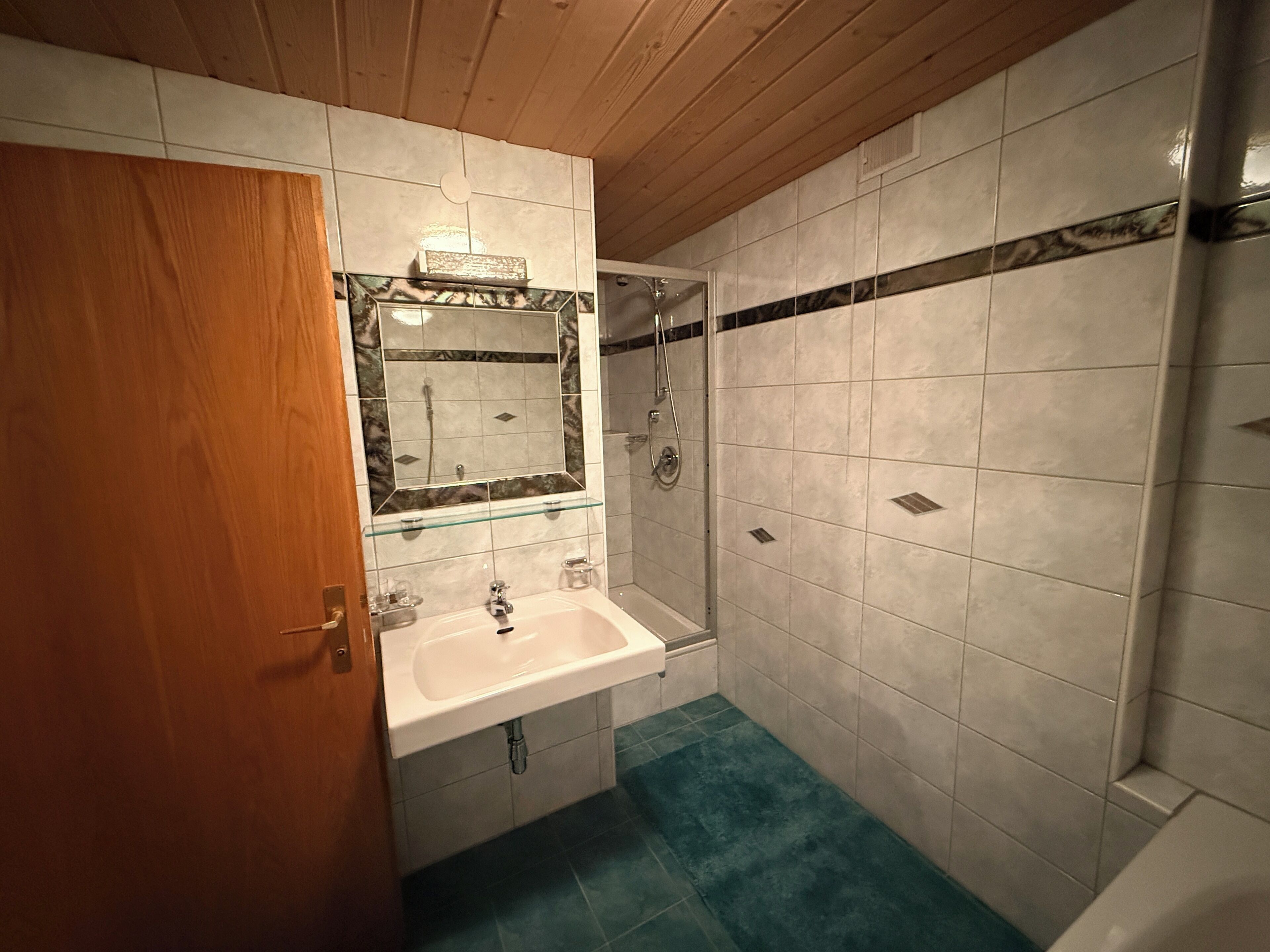 Ein Badezimmer mit Holzboden, weißen Fliesen, einer Glasablage über dem Waschbecken und einer Duschkabine.