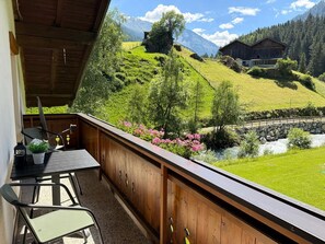 A balcony with a table and chairs, overlooking a green valley with a river and a mountain in the background. - Cozy vacation apartment for 4 people in the Pitztal Valley—quiet and close to nature (Jerzens)