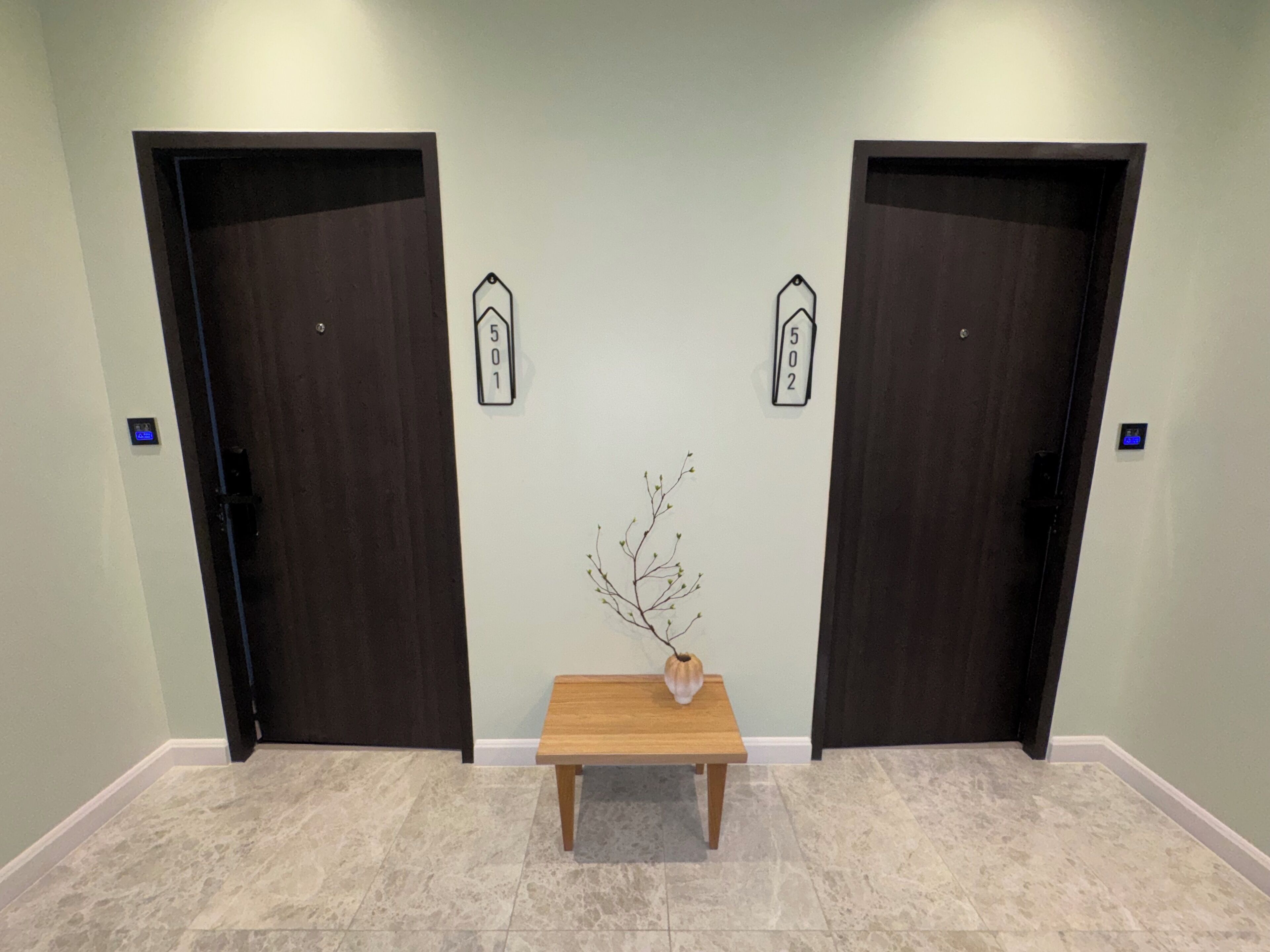 Un pasillo con dos puertas de madera oscura, una pequeña mesa de madera con un florero y un letrero con un número colgado en la pared.