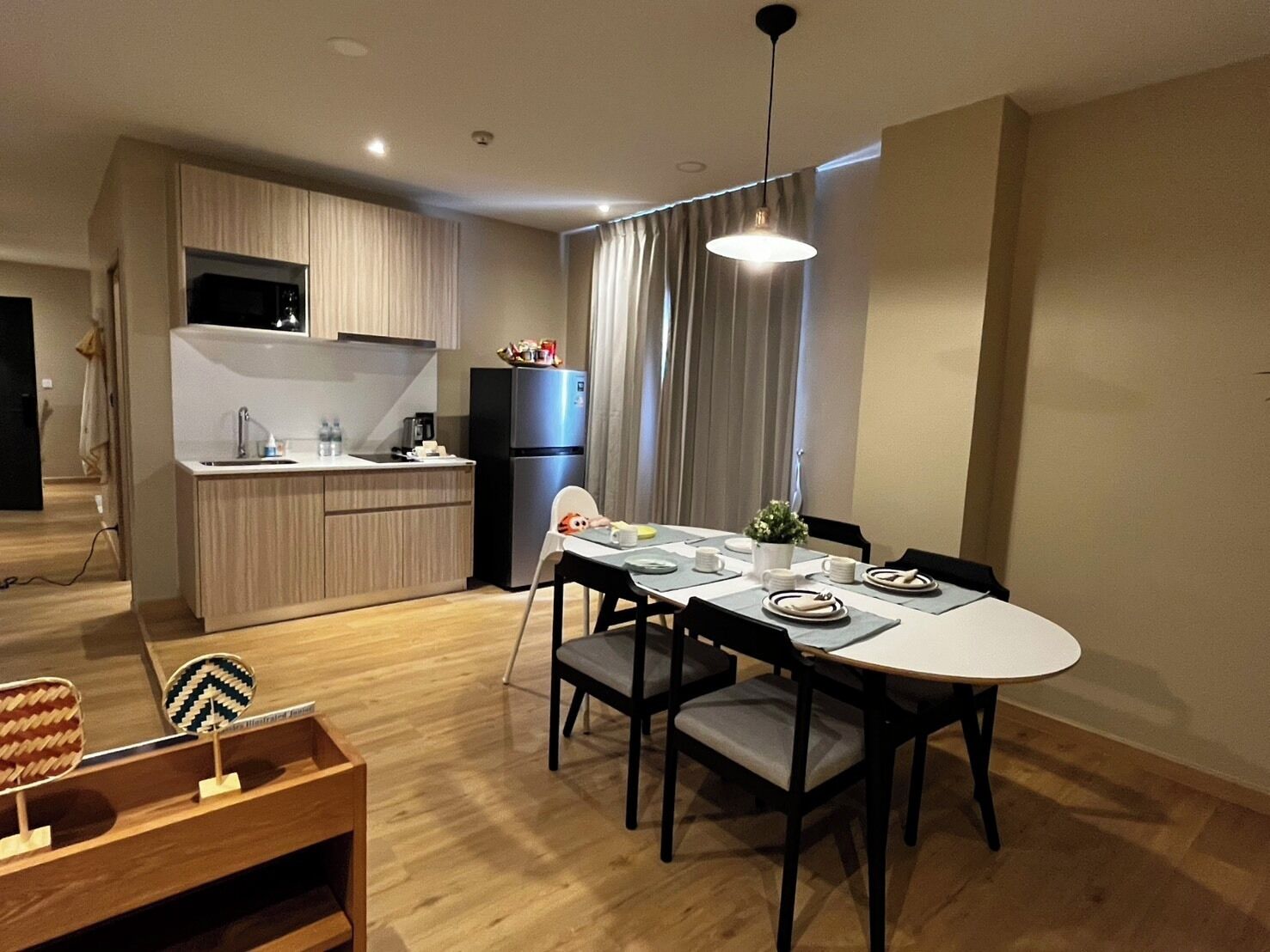 Een moderne keuken met een inbouwoven en koelkast, een eethoek met een ronde tafel en stoelen, en een klein zithoekje met een fauteuil en bijzettafeltje.