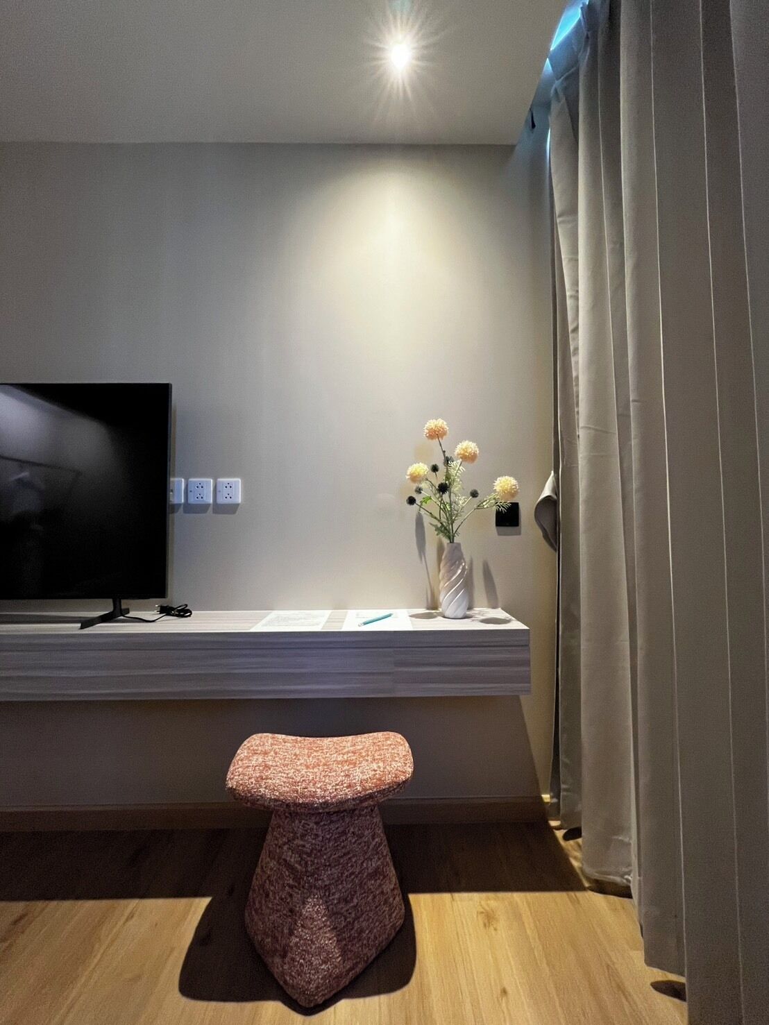 Una sala de estar moderna con un televisor de pantalla plana, una consola blanca, un florero con flores, un taburete texturizado y una cortina.