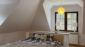 A bedroom with a sloped ceiling, wooden beams, a bed with a plaid bedspread, two bedside lamps, and a window with a view of trees.