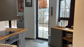 A modern kitchen with a central island, a built-in oven, and a sink. There is a window with a view of trees and a door leading outside.