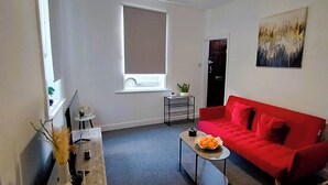 A compact living space with a red sofa, a small table with a bowl of fruit, a potted plant, and a painting on the wall. - Prime Town Centre-Location-Station-Airport-WiFi-Parking (Luton)